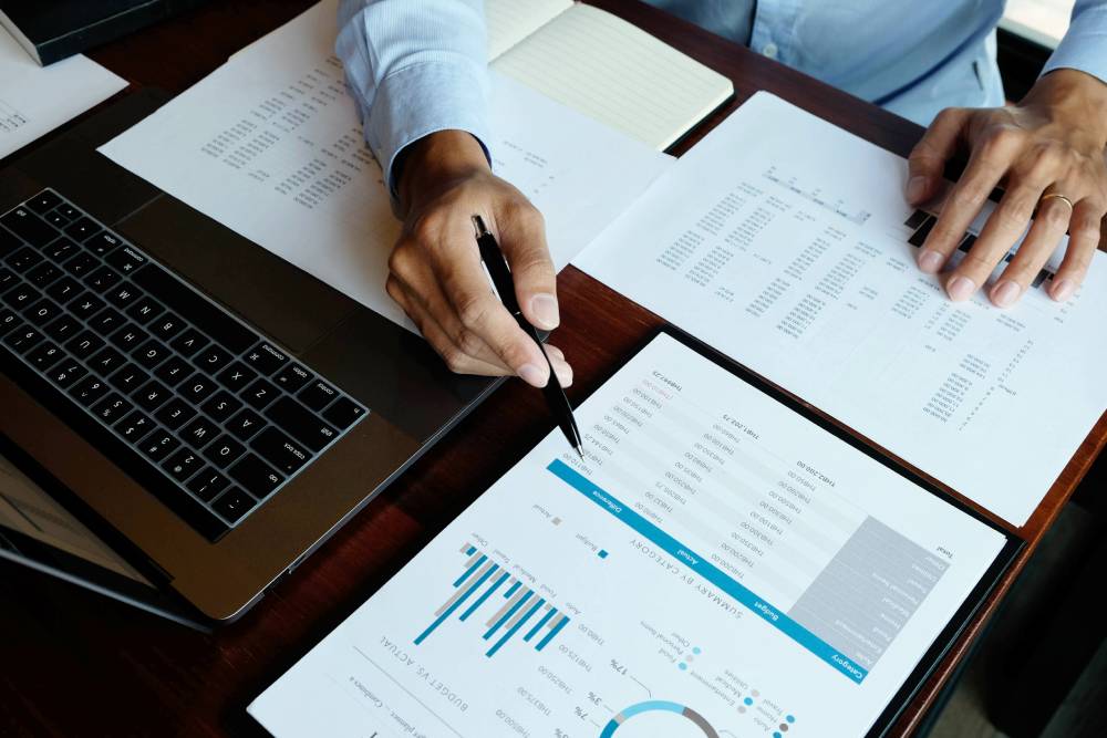 Hands with a pen reviewing printed financial reports beside a laptop