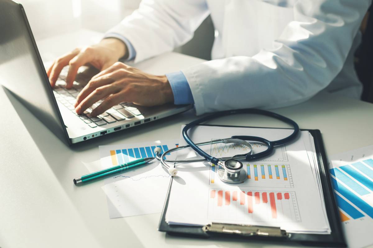 5 Financial Mistakes Physicians Make (And How to Help Avoid Them) 2 Doctor typing on a laptop beside a stethoscope and financial charts