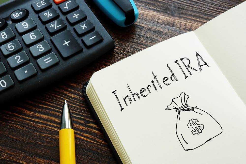 Notebook page reading “Inherited IRA” with a drawn money bag, beside a calculator and pen