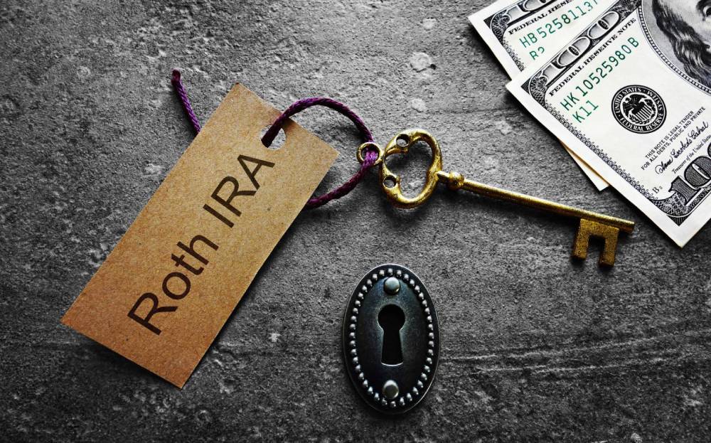 Paper tag reading “Roth IRA” tied to a gold key, beside a keyhole and dollar bills